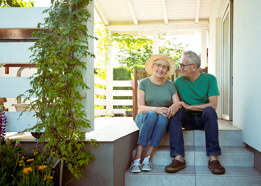 Smiling retired, senior couple posing in front of their house, representing positivity throughout shifts in the housing market.