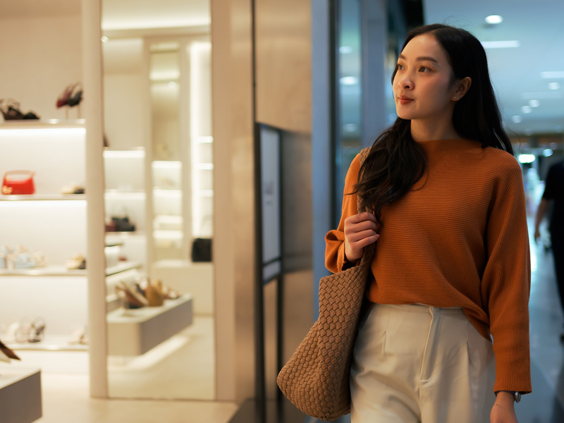 An young asian women looking at shoes and purses in a mall feeling recent consumer concerns with inflation and recent stock changes.